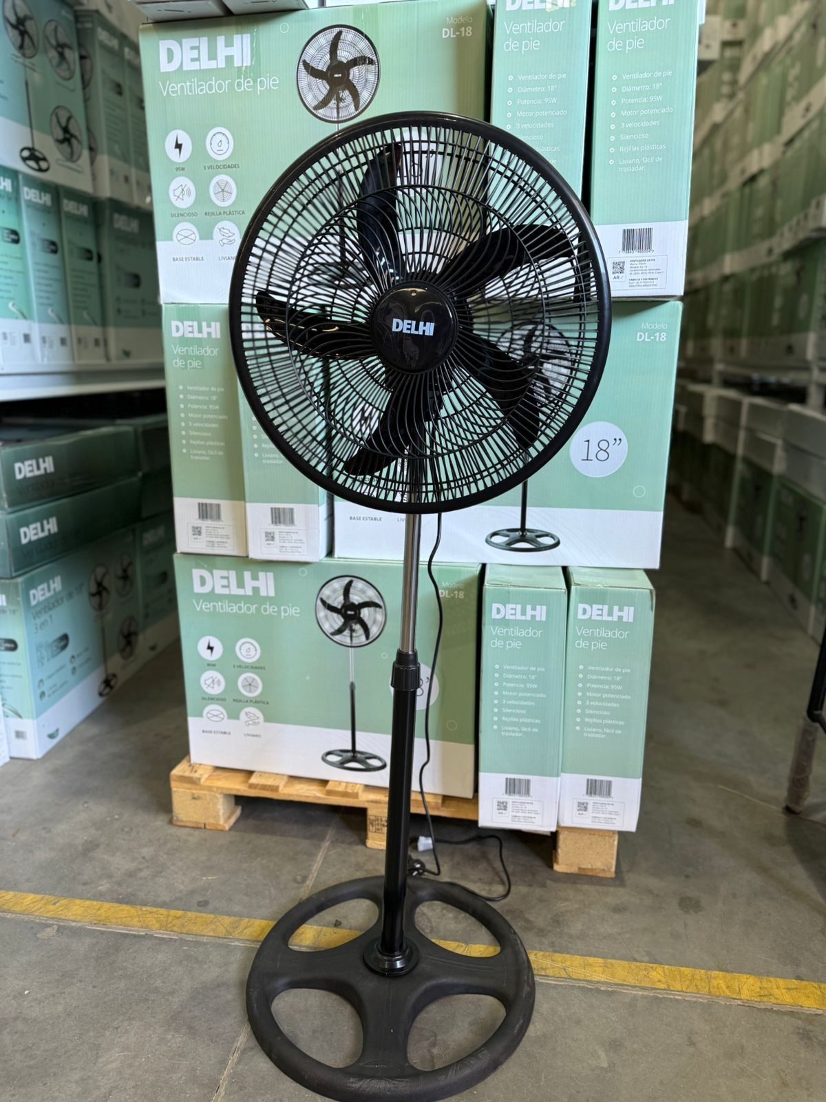 Ventilador de pie de color negro con base circular, delante de una paleta de ventiladores en forma de caja.