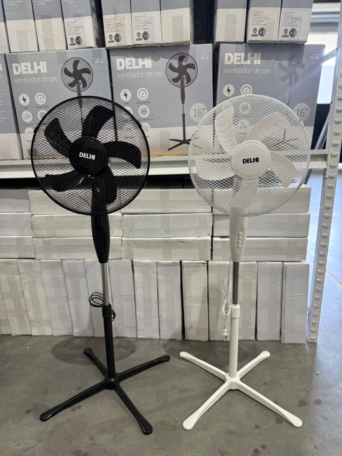 Dos ventiladores de pedestal, en blanco y negro, delante de una pila de ventiladores en caja.