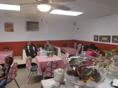 Diners sit at tables covered with red-and-white checkered cloths in a cafe with a display of donuts in the foreground.