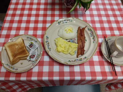 A breakfast with scrambled eggs, bacon, a scoop of white food, and two slices of toast on a red-and-white checkered cloth.