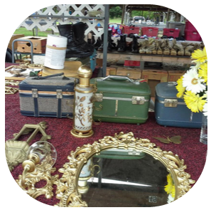 A table at a flea market display holding vintage train cases, an ornate gold mirror, a floral lamp, and yellow flowers.