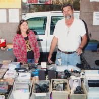 Two people stand behind a table filled with assorted electronic goods and containers at an outdoor market.