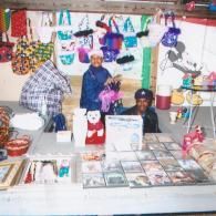 Two people sit at a table covered with goods like small bags, a toy bear, and packaged items in a shop-like setting.