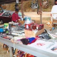 A vendor stands behind a table filled with assorted goods, including boxes, bags, and apparel, at an outdoor market.