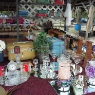 A flea market table displays vintage items, including a yellow suitcase, a blue cooler, glassware, and decorative accents.