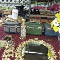 A flea market table displays three vintage suitcases, a glass bottle, a gold ornate mirror, and a vase of white flowers.