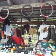 A stall at an outdoor market displays assorted clothing, hats, and small items hanging under a wooden roof structure.