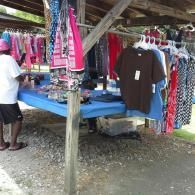 A person stands at a blue table covered in clothes for sale under a wooden outdoor shelter.