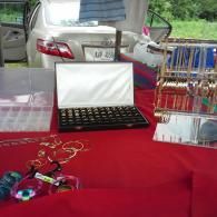 A vendor display on a red table featuring a ring case and a necklace stand in front of a parked silver car.