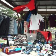 A market stall under an open-sided roof displaying rows of clothing, sunglasses, and assorted merchandise on tables.