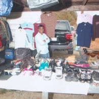 A person stands behind a table displaying clothes and shoes at an outdoor market with a black vehicle in the background.