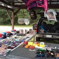 A roadside vendor stall under a covered structure displays various items, including bags, clothing, sandals, and eyewear.