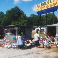 People browse through boxes of items and goods for sale at an outdoor flea market under a sunny, blue sky.