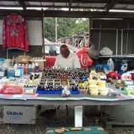 A vendor sits behind a table at an outdoor market, displaying rows of small bottles, containers, and a hanging red jacket.