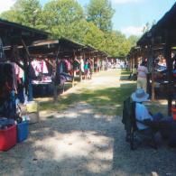 Open-air market with clothing racks under wooden pavilions, grass pathway, and a seated person in the foreground.