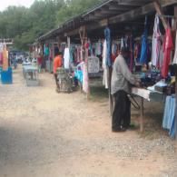 A vendor stands at an outdoor market stall displaying rows of hanging garments on a dirt lot.