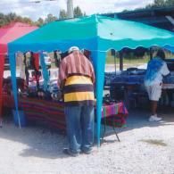 Two people stand under a teal canopy at an outdoor market, browsing items displayed on a table.
