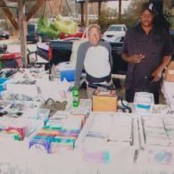 Two individuals stand behind a folding table covered with various items for sale at an outdoor market.