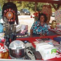 A smiling person behind a vendor booth displaying motorcycle gear, a helmet, and white boxed items under a canopy.