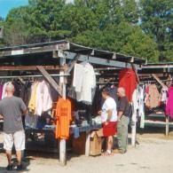 People browsing racks of clothing under a covered outdoor shelter at a market.