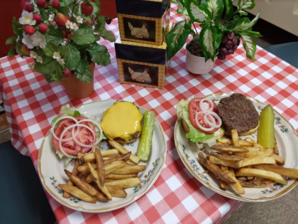 Two plates of cheeseburgers and fries served on a red and white checkered tablecloth with two small potted plants.
