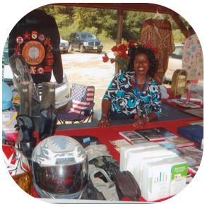 A smiling person sits at a booth featuring a motorcycle helmet, boots, books, and a garment with patches, outdoors.