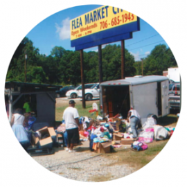 People sort through items displayed from an open trailer at an outdoor flea market under a blue signage board.