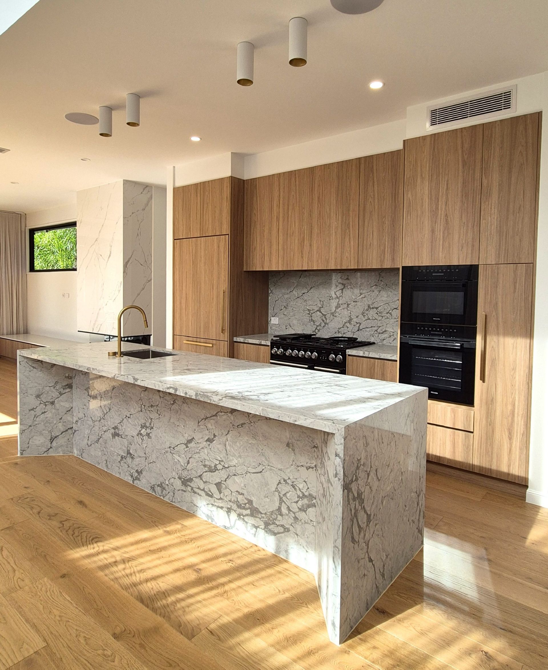 Modern kitchen with wood cabinets, marble island, black appliances, and sunlit hardwood floors.