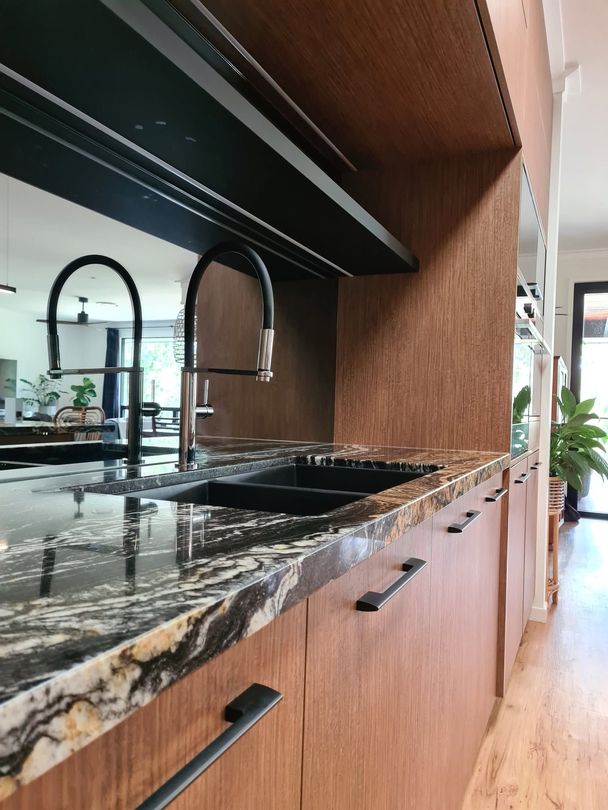 Modern kitchen counter with black marble, wood cabinets, and double black faucets by a bright window