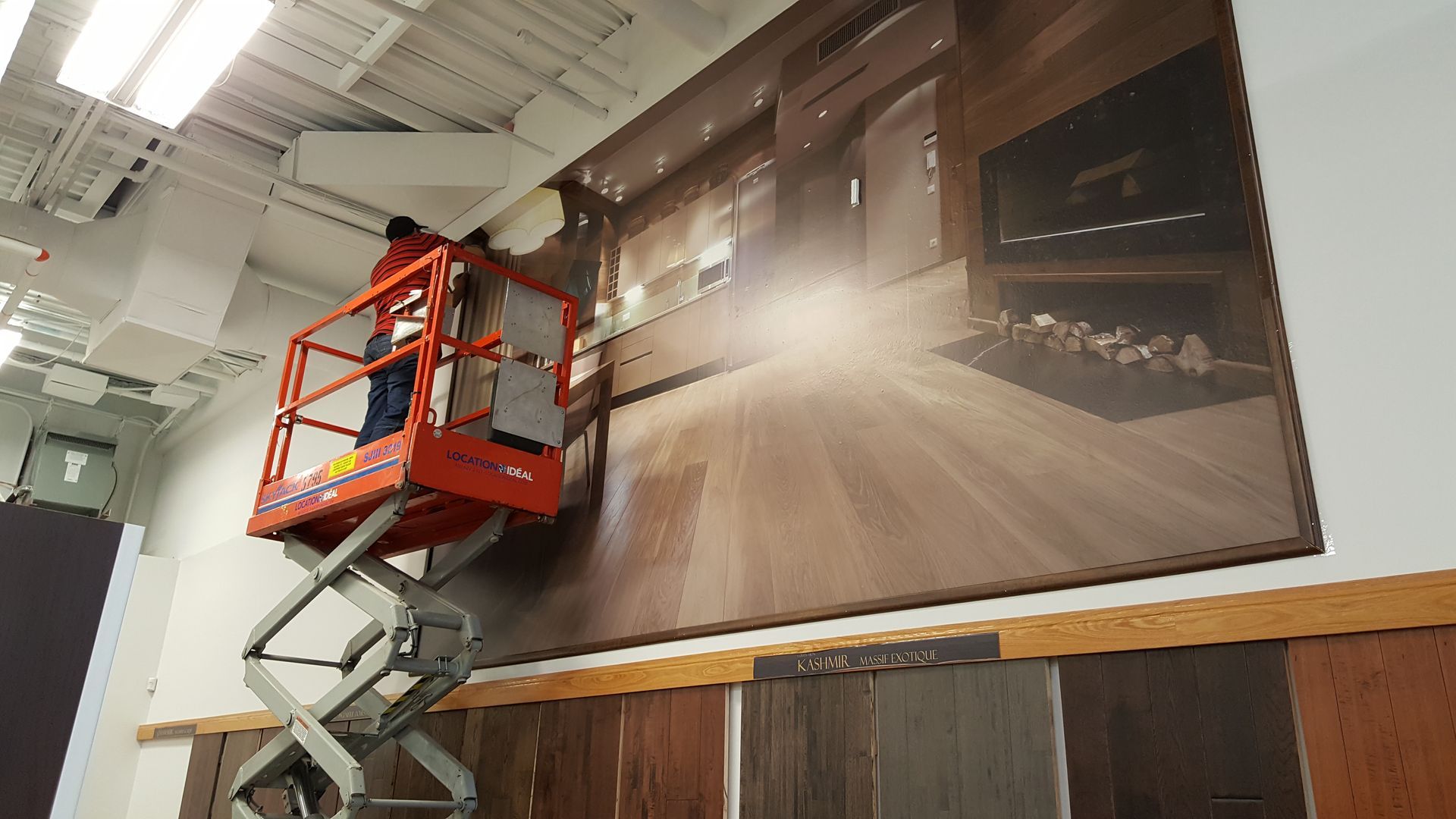 Two workers on a lift installing a large graphic on a wall, interior setting.