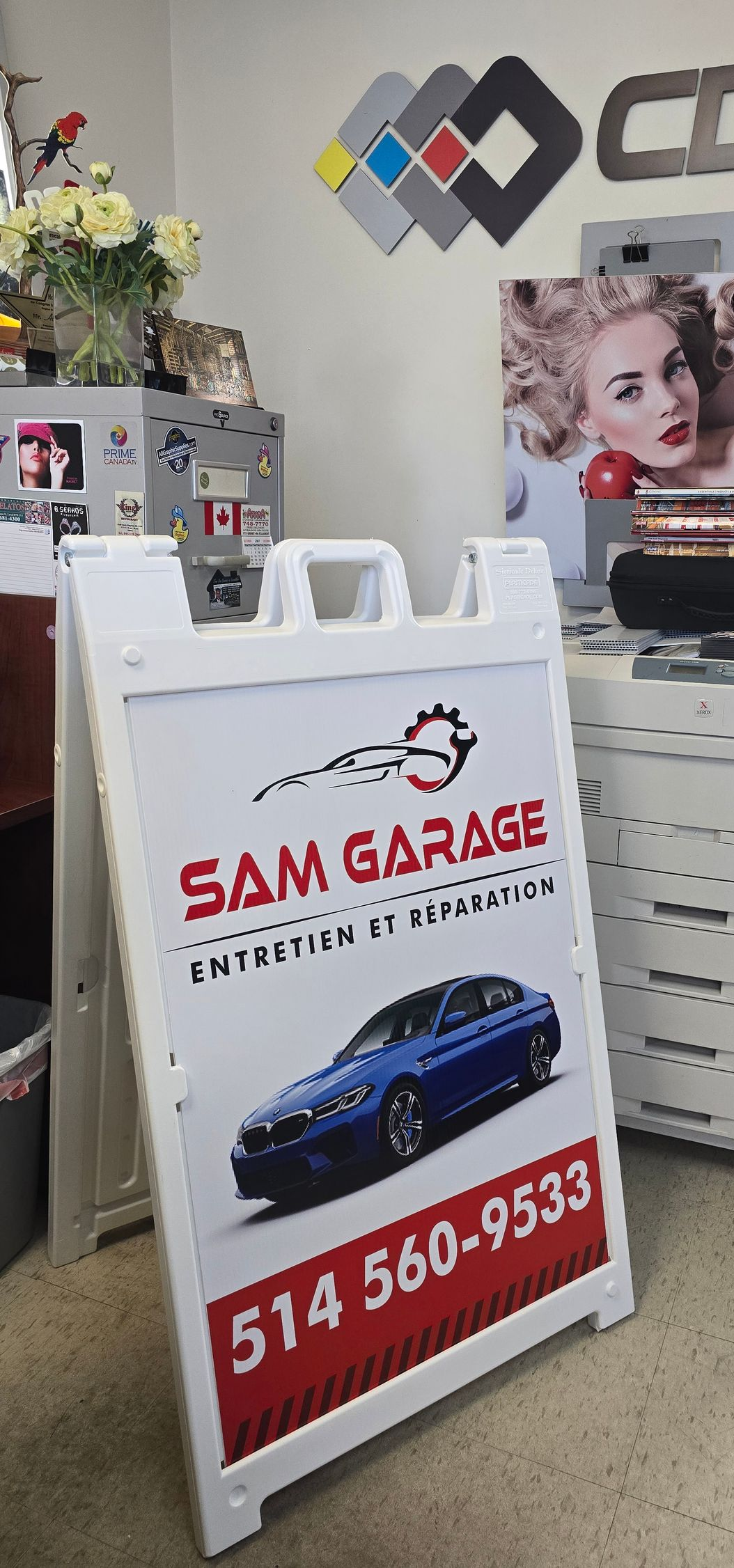 White A-frame sign for Sam Garage with car image, phone number, and text in a shop.