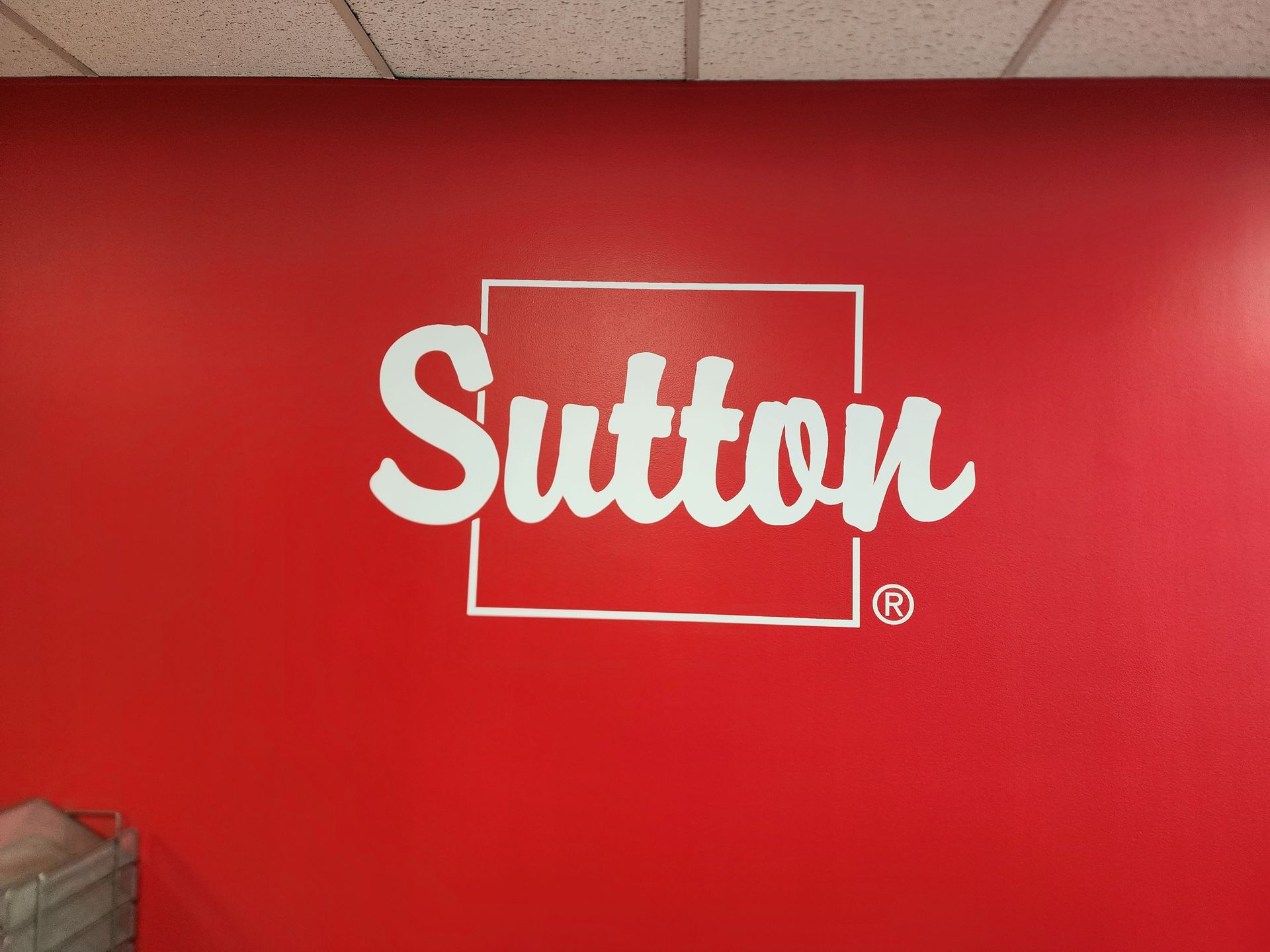 Logo blanc « Sutton » dans un carré sur un mur rouge.
