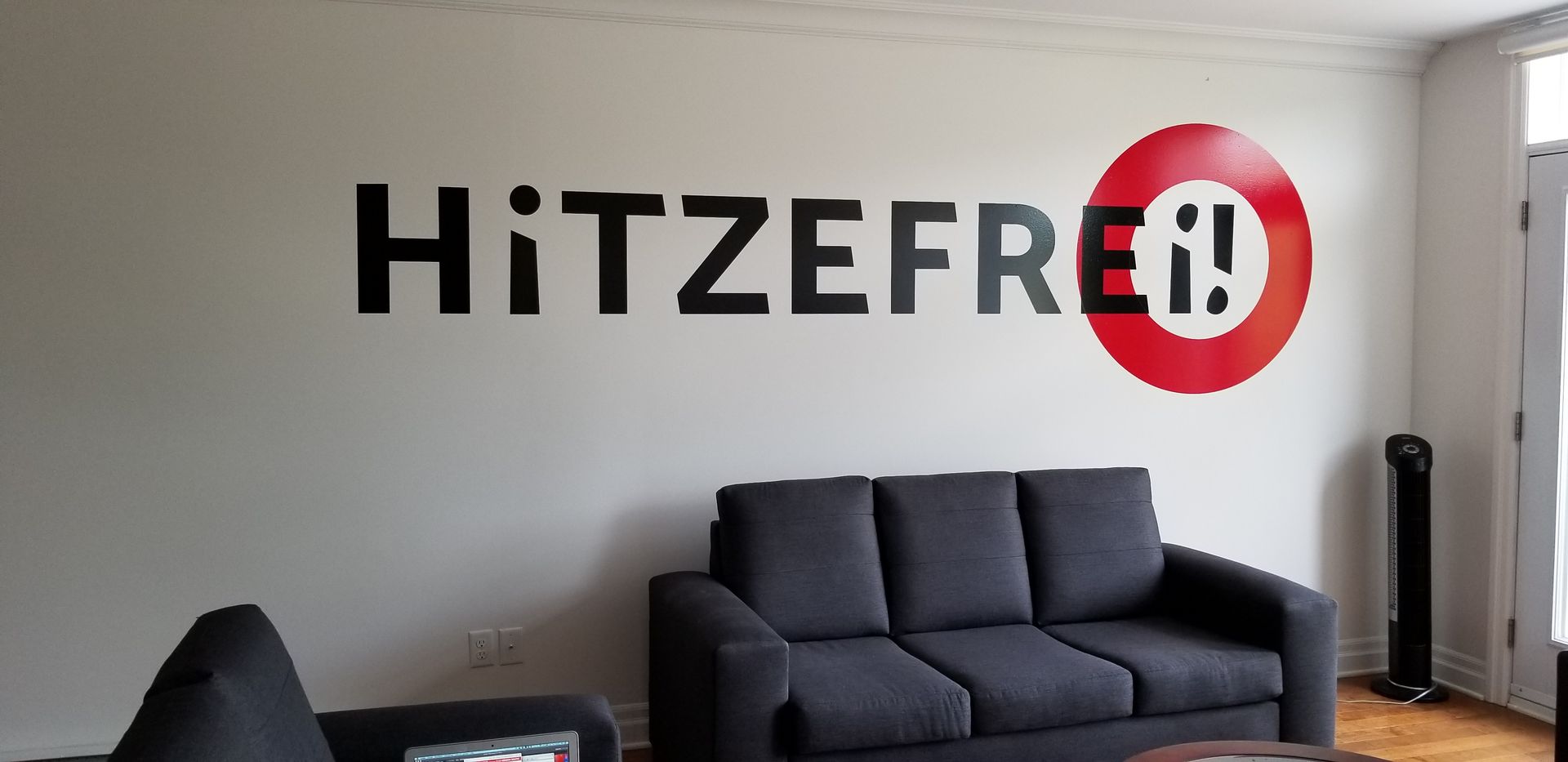 Une pièce avec un canapé gris foncé, les mots « HiTZEFREi! » sur le mur blanc, avec un logo en forme de cercle rouge.
