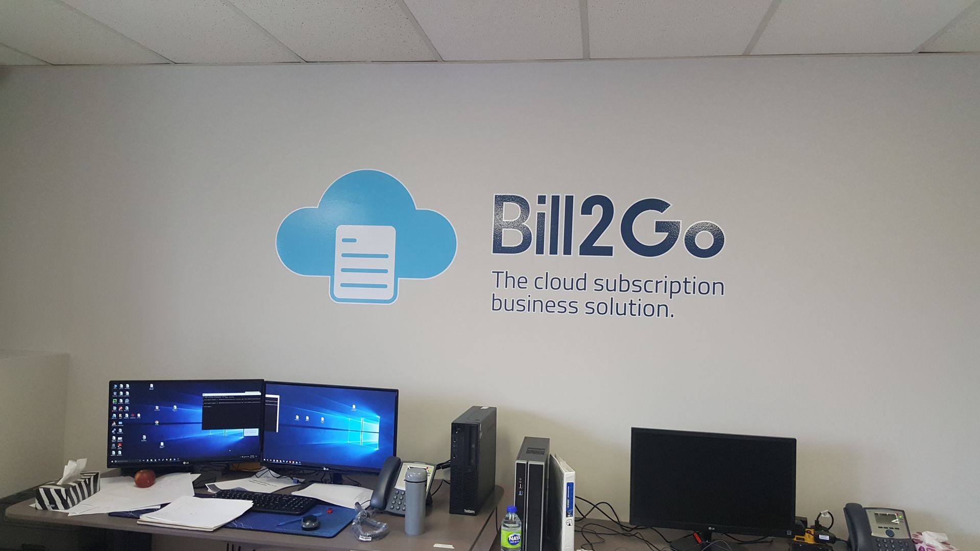 Bureau avec le logo « Bill2Go », deux écrans, un ordinateur et des articles de bureau.