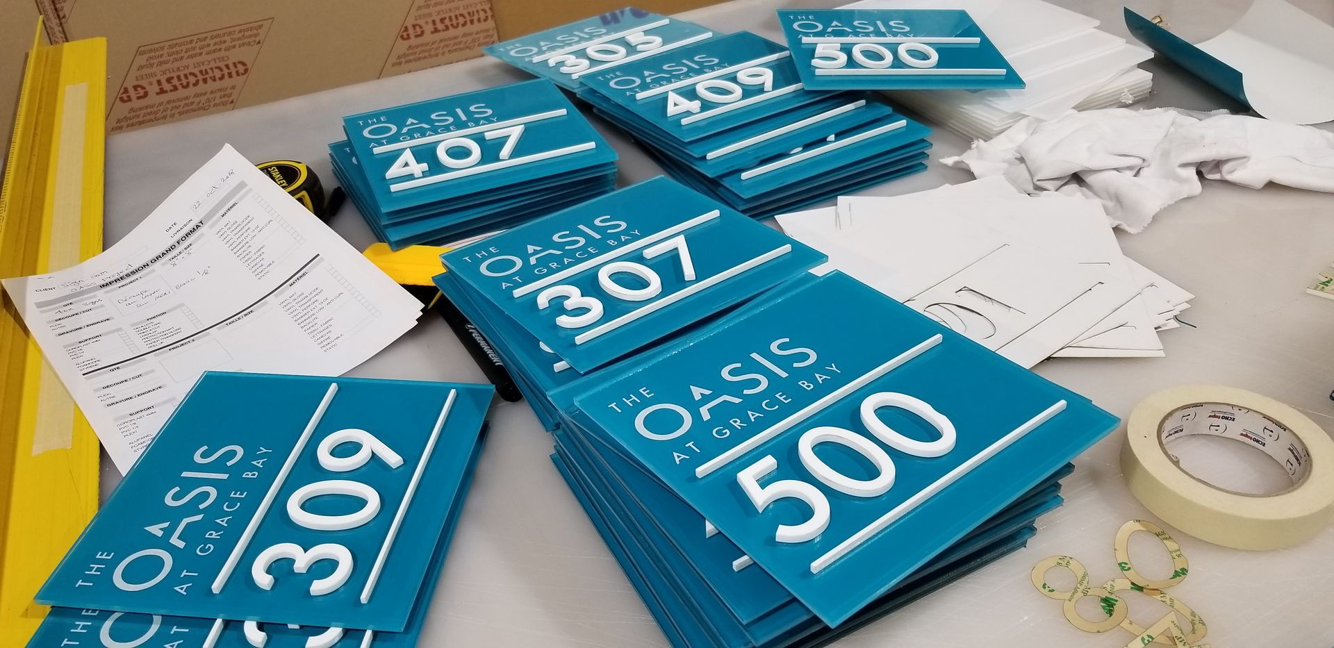 Piles de panneaux bleu sarcelle avec du texte blanc, étiquetés « L'Oasis » et des numéros d'appartement.