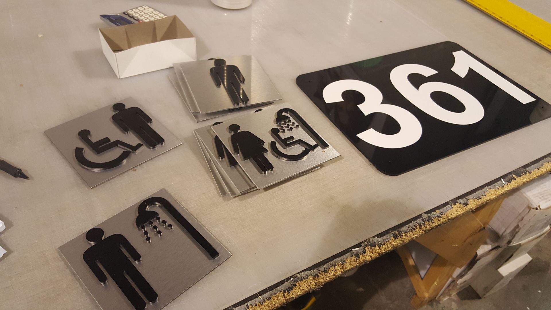 Signalétique avec toilettes, douche et numéro de bâtiment 361. Symboles noirs et texte blanc sur acier inoxydable.