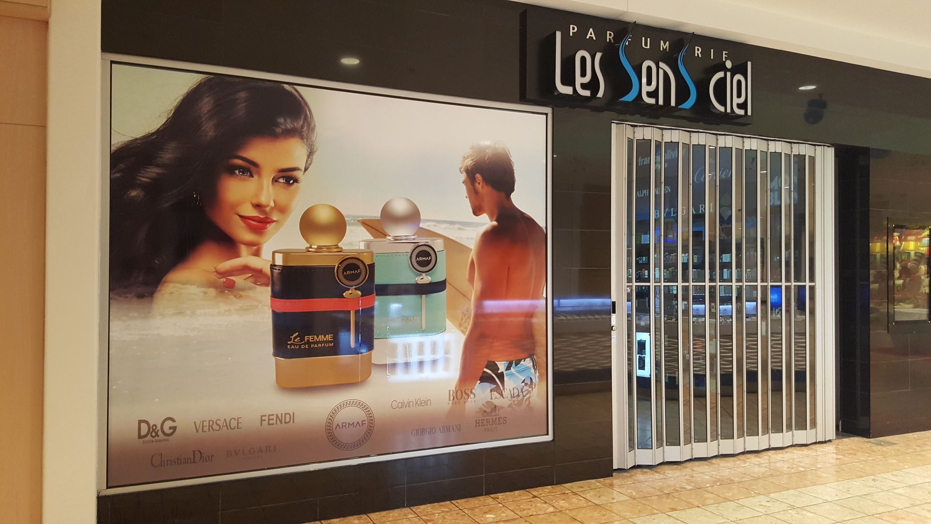 Une parfumerie, « Les Sens Ciel », avec une femme et un homme sur une bannière. Portail de sécurité métallique fermé.
