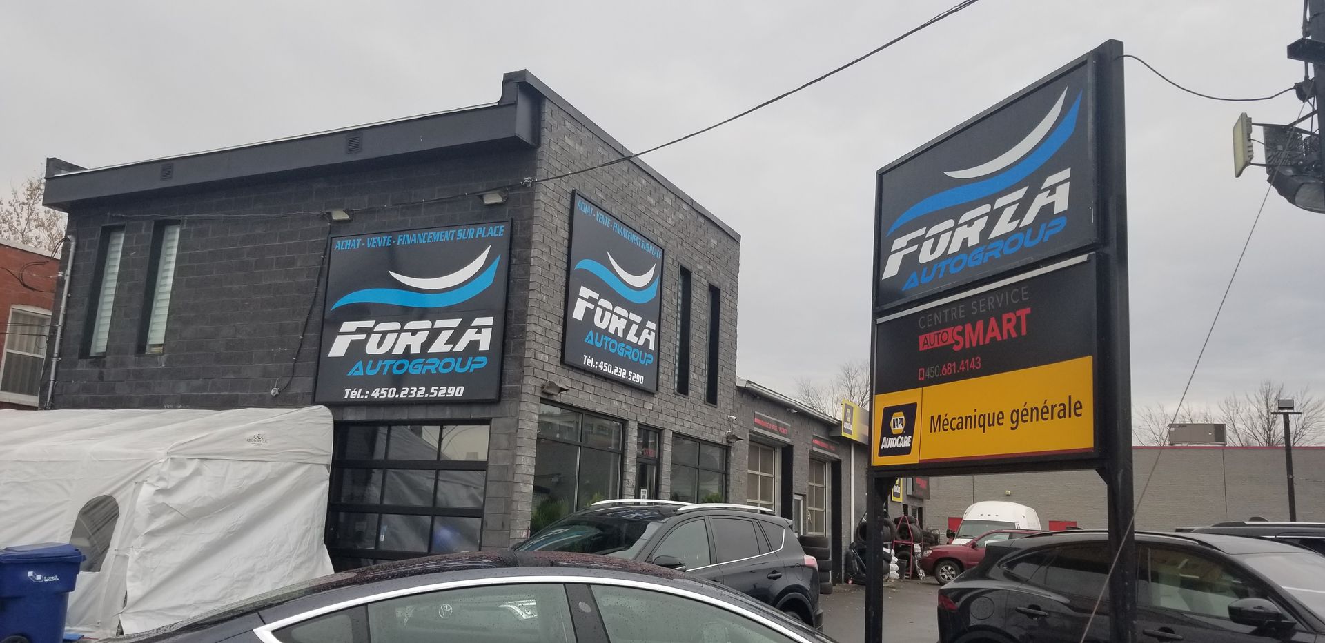 Bâtiment du groupe Forza Auto avec signalisation, voitures devant et ciel couvert.