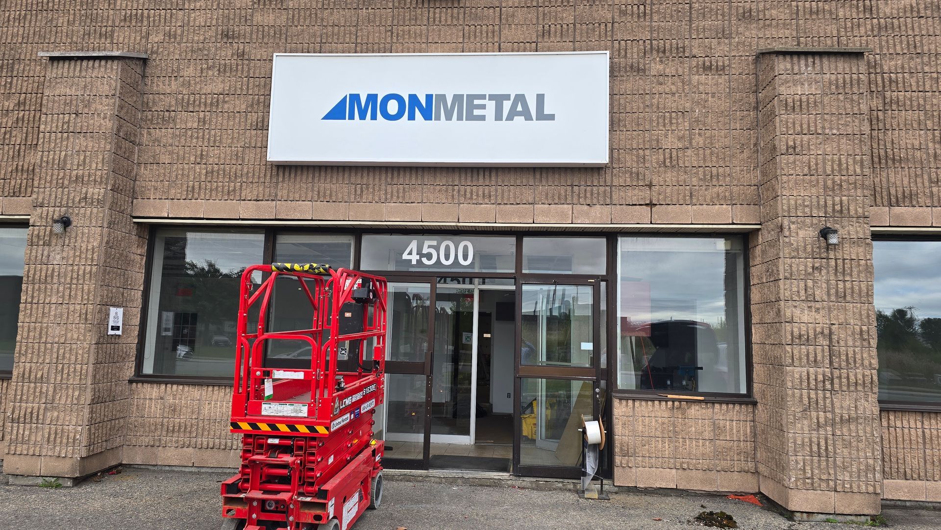 Entrée du bâtiment MONMETAL avec signalétique. Un ascenseur rouge se trouve devant les portes vitrées. L'adresse, 4500, est indiquée au-dessus de l'entrée.