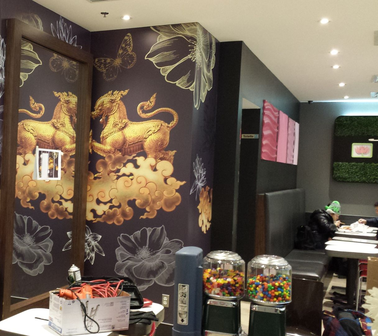Intérieur de restaurant avec une fresque murale représentant un lion doré sur un mur violet foncé, des distributeurs de boules de gomme et une personne assise à une table.