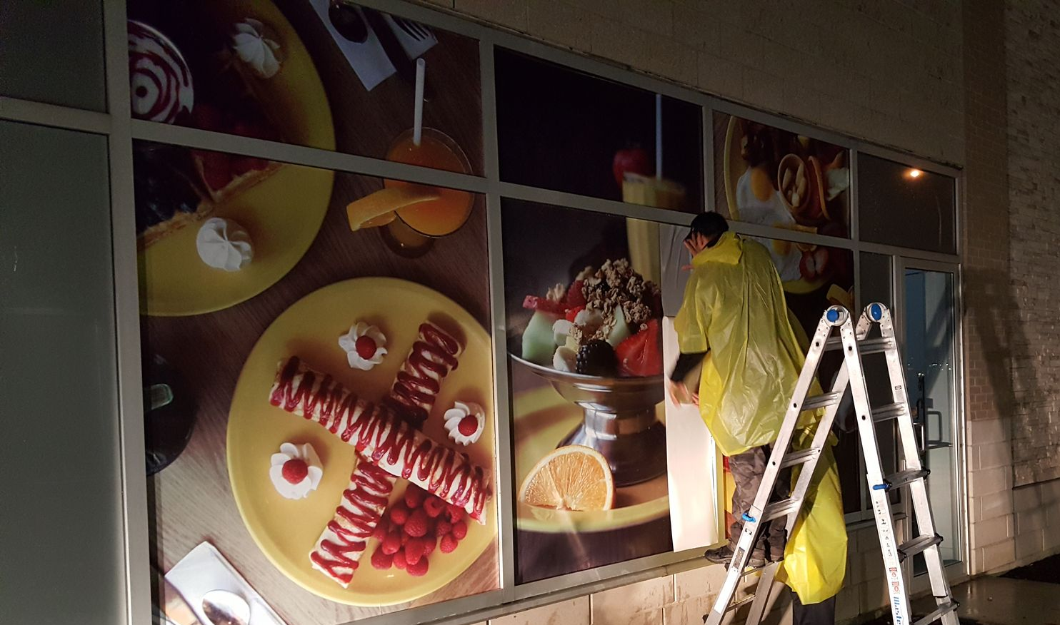 Une personne en imperméable jaune sur une échelle installe une publicité alimentaire sur la vitrine d'un magasin.