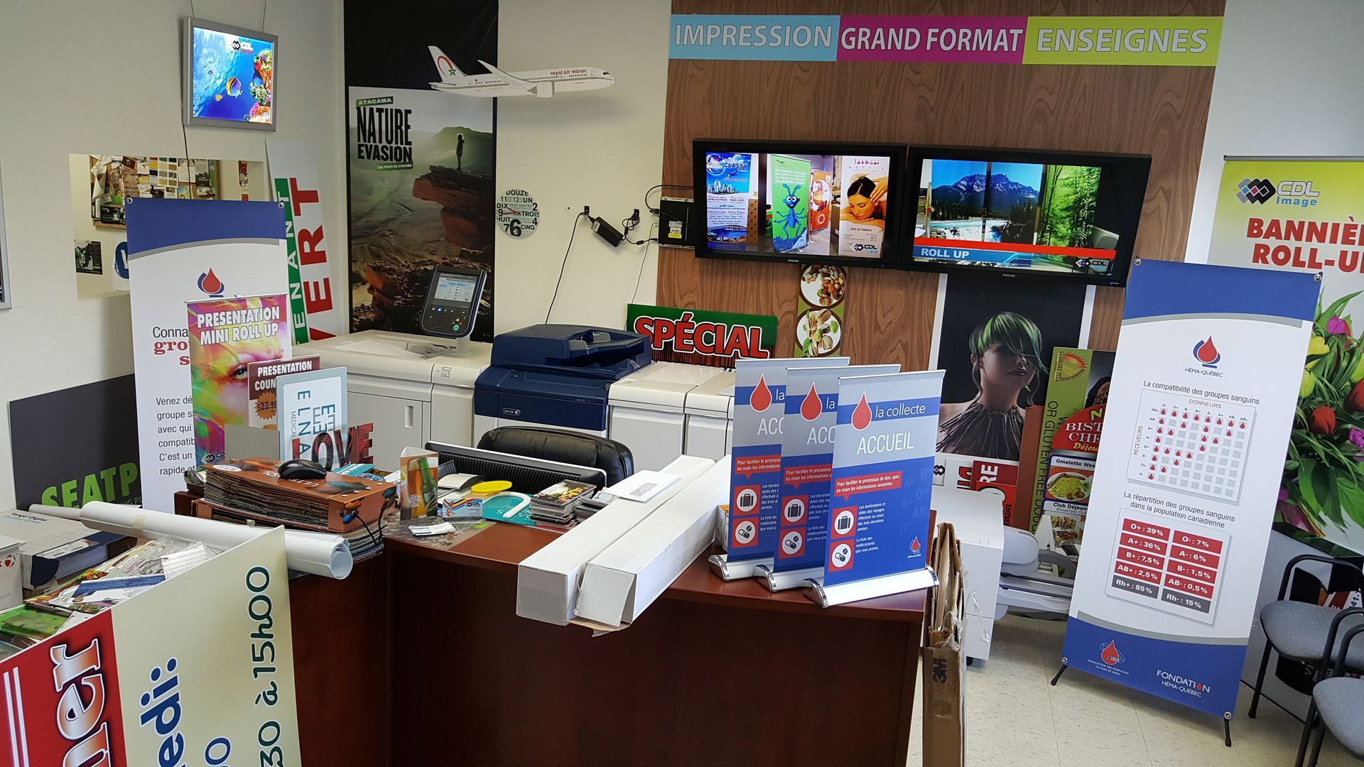 A cluttered office with displays. Blue and white promotional materials are prominent. Three screens show various images.