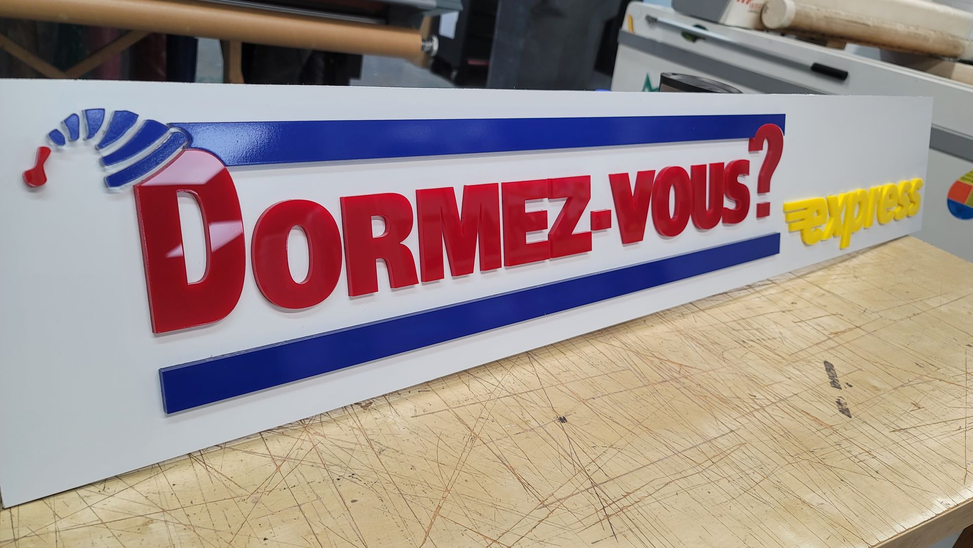 Panneau blanc avec inscription « DORMEZ-VOUS ? » en lettres rouges, lignes bleues et bonnet de nuit. Logo jaune et or à droite.