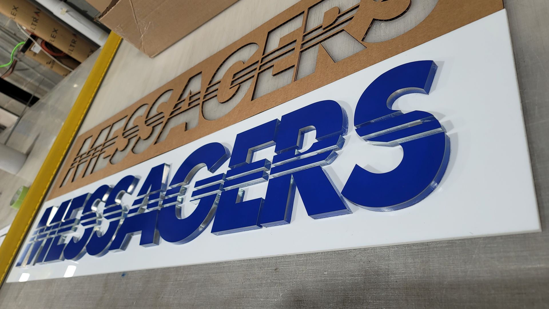 Un panneau qui écrit « MESSAGERS » en lettres bleues superposées sur fond blanc, avec un pochoir en carton au-dessus.