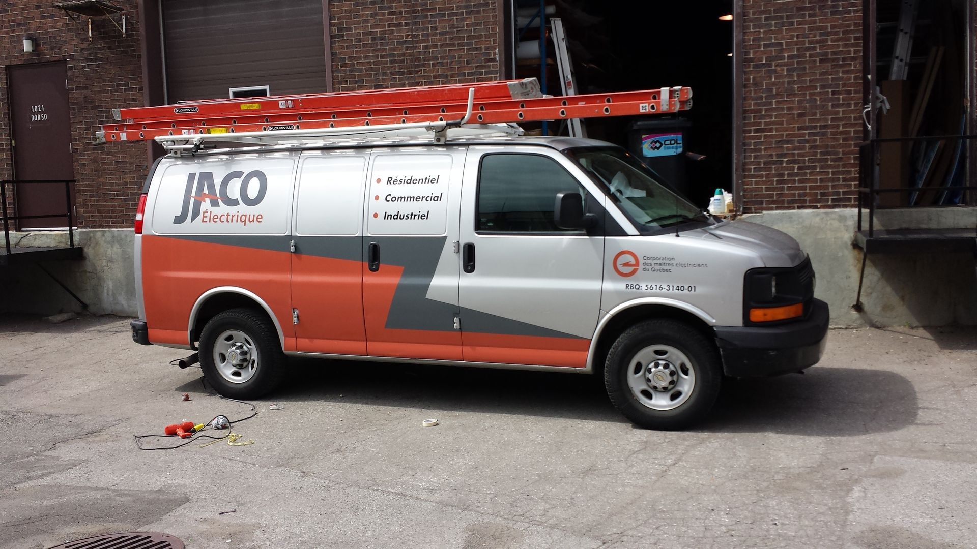 Une camionnette JACO Services avec une échelle sur le dessus, garée près d'un bâtiment en briques.