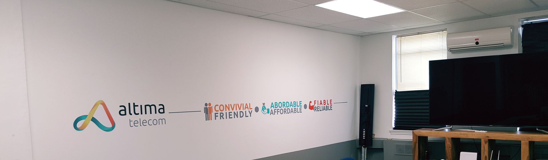 A white-walled room with a logo on the wall, two large black screens on a wooden table and a bright window.