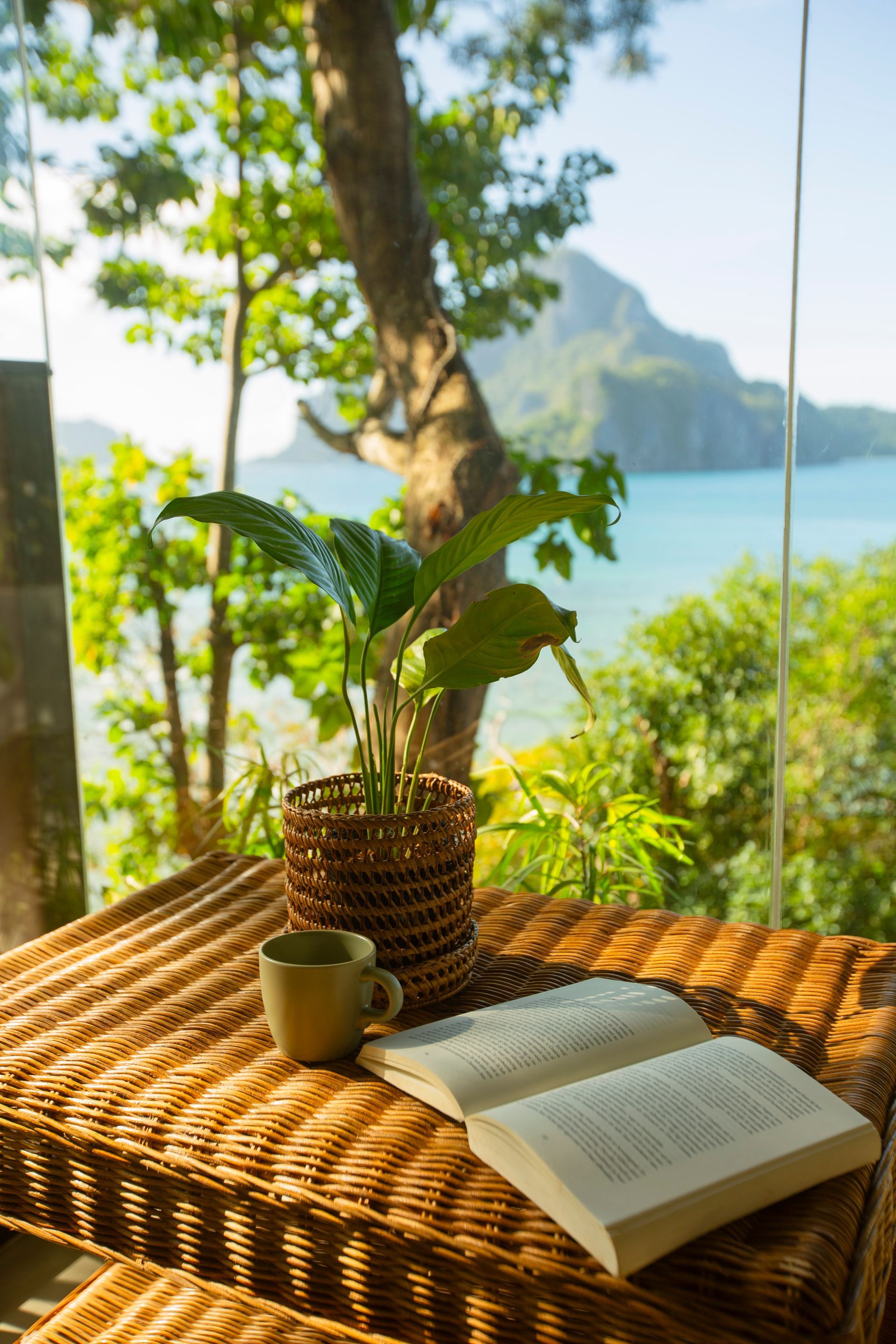 A book and a cup of coffee are on a wicker table with a view of the ocean.