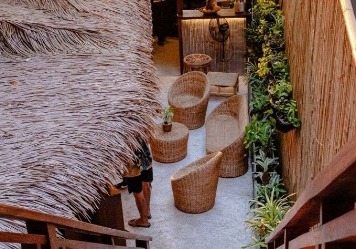 A person is standing under a thatched roof next to wicker chairs and tables.