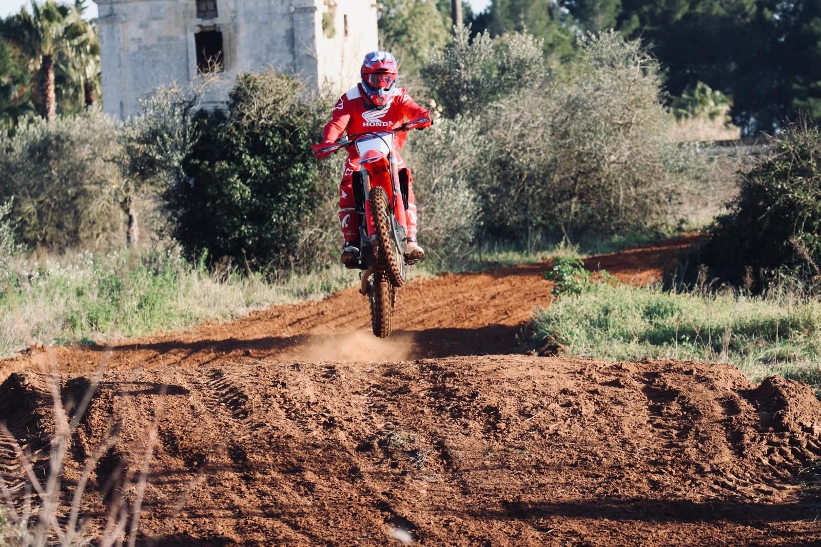 Motociclisti che sfrecciano su una strada sterrata, sollevando polvere in una foresta con le montagne sullo sfondo.