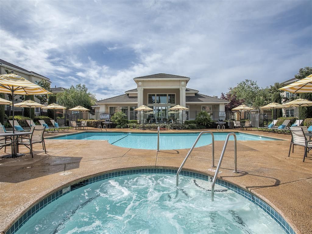 Outdoor community pool area with lounge chairs, umbrellas, and a clubhouse in the background.
